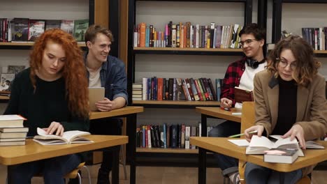 intelligent female and male students learning, preparing for exams in college library sitting by the separate tables over bookshelf background. charming young people learning at library