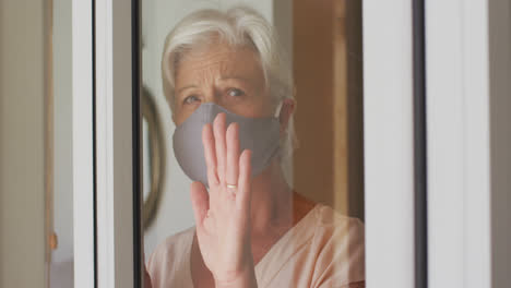 Mujer-Caucásica-Mayor-Con-Mascarilla-Mirando-Por-La-Ventana-En-Casa