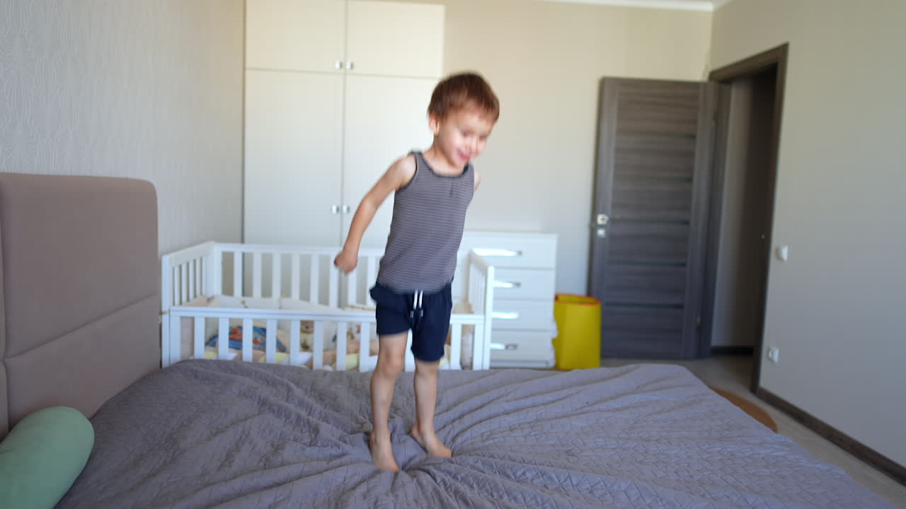 Energetic baby boy jumps merrily on the big bed in the bedroom. Kid lies down and looks up.