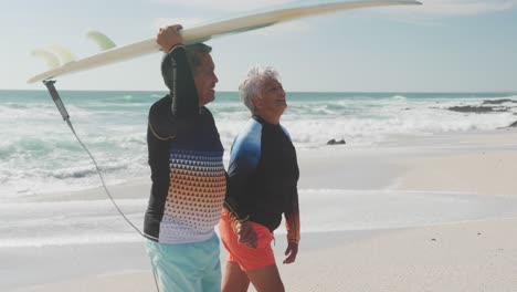 side view of happy senior hispanic couple walking on beach with surfboard