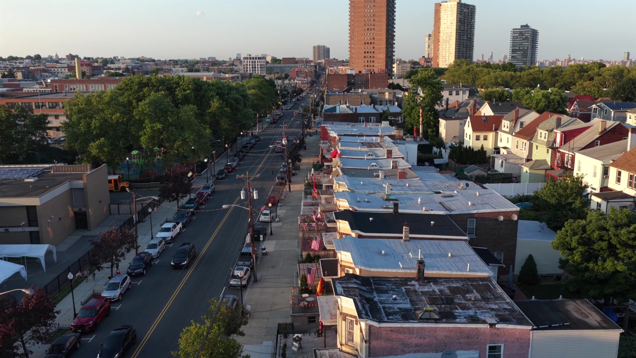 Tetti di case a schiera con bandiere americane sui balconi fiancheggiano una strada alberata, alte torri di appartamenti si ergono oltre file di auto parcheggiate al tramonto dorato a Union City, New Jersey. Ampia ripresa aerea con drone