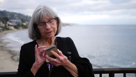 Una-Mujer-De-Mediana-Edad-Con-Cabello-Blanco-Y-Anteojos-Enviando-Mensajes-De-Texto-En-Su-Teléfono-De-Vacaciones-En-Laguna-Beach,-California-En-Cámara-Lenta