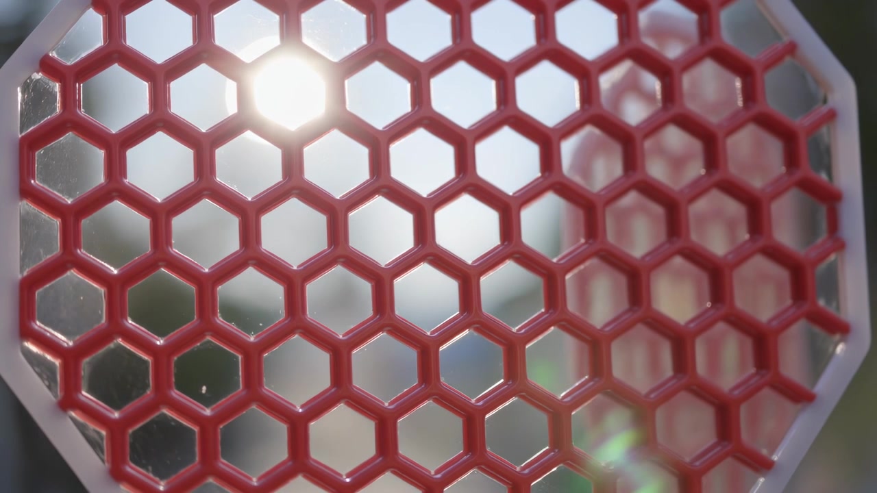 Sol filtrándose a través de una malla hexagonal roja con un marco octogonal blanco, causando un destello de lente verde en el exterior.
