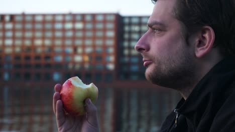 close up of a young man eating a red apple and looking into the distance on a pier during the evening