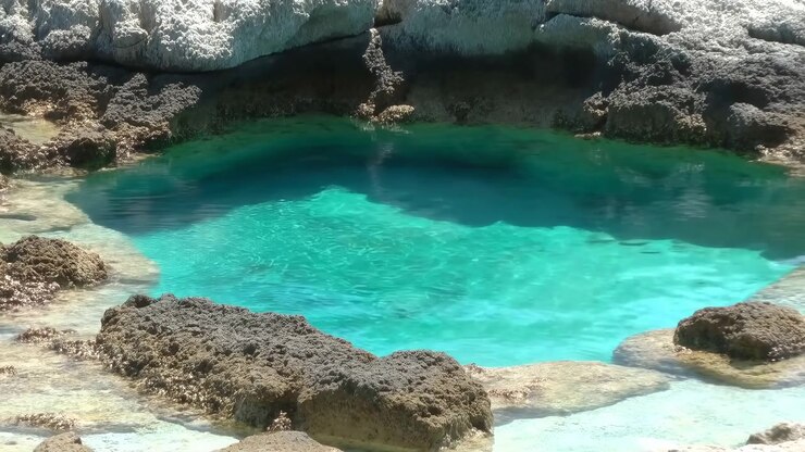Clear blue-green water shimmers under bright sunlight as small surface ripples move across a rocky basin, with pale shallows, dark submerged areas, and rough textured stone.