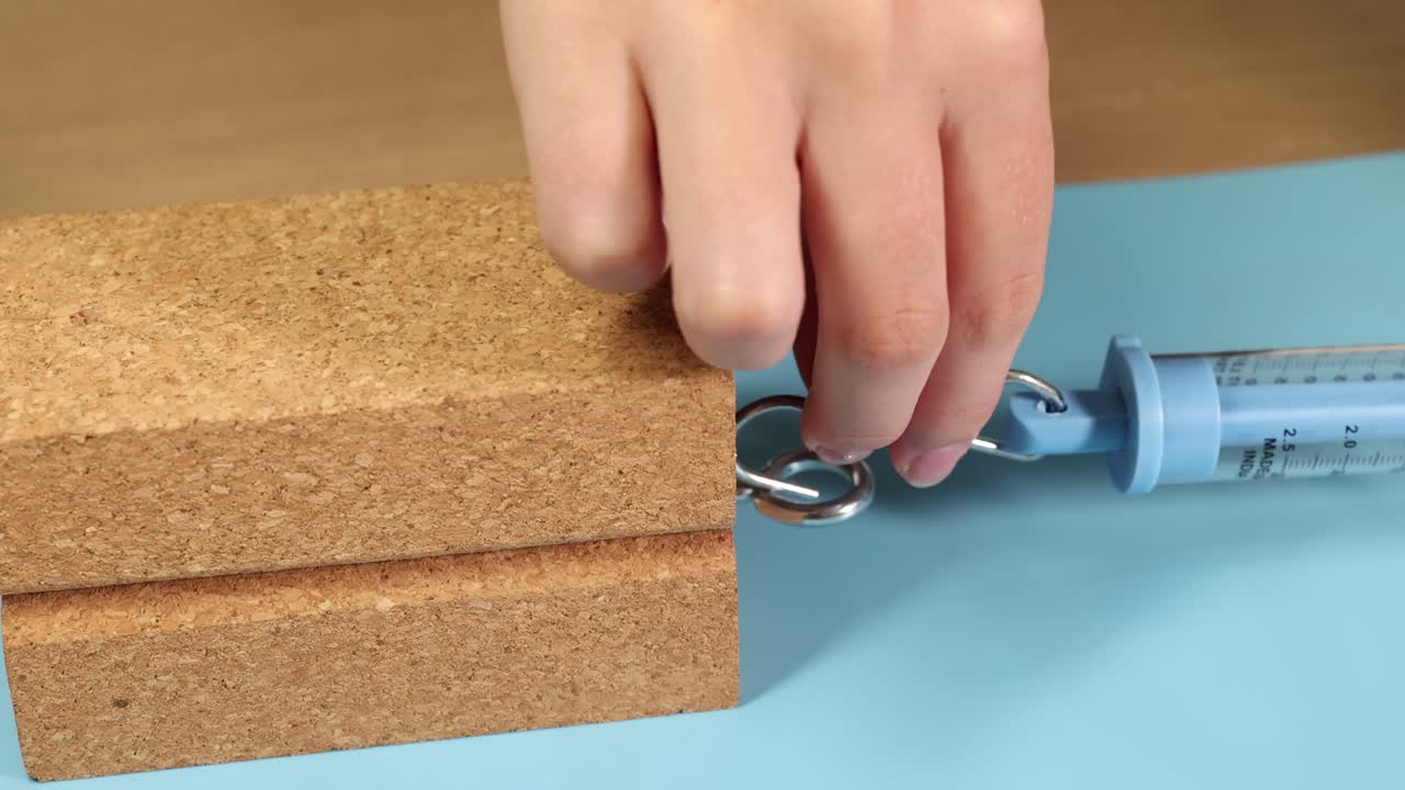 A spring scale measures friction as a hand pulls a wooden block across a surface, highlighting force dynamics
