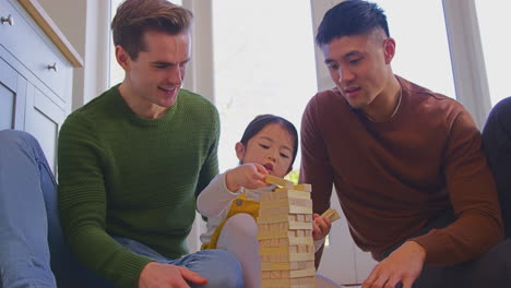 family with two dads playing game with daughter at home stacking wooden bricks into tower