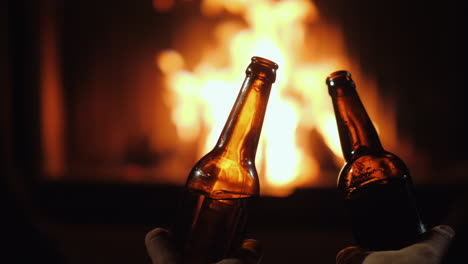 Dos-Botellas-De-Cerveza-En-Manos-De-Los-Hombres-Sobre-Un-Fondo-De-Fuego-En-Una-Chimenea-Primer-Plano