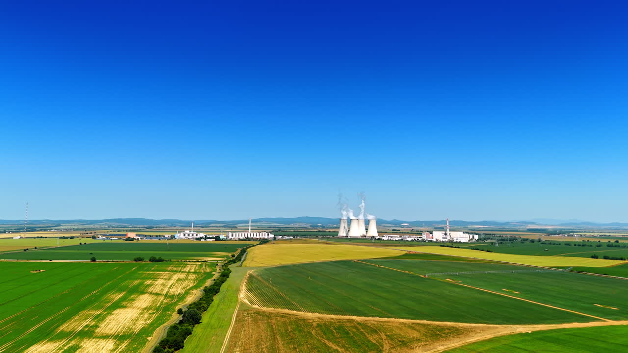 Going up over the green countryside fields. Modern plant works in the rural area producing white smoke