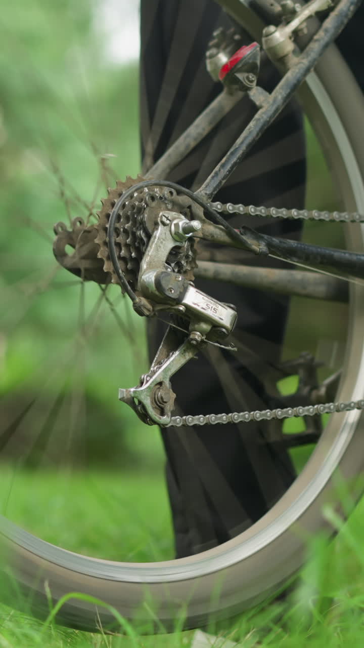 primer plano de un individuo con pantalones y zapatillas negras de pie junto a una bicicleta estacionada en un campo cubierto de hierba, levantando la rueda trasera y pedaleando brevemente, haciendo que la rueda gire con un movimiento borroso visible