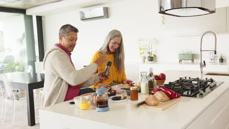 breakfast, multiracial senior couple sitting at kitchen island with coffee and pastries, at home