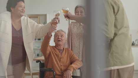 steadicam of happy african woman, handicapped caucasian man in wheelchair, grey-haired asian lady and unrecognizable man standing in apartment, clinking glasses and drinking white wine