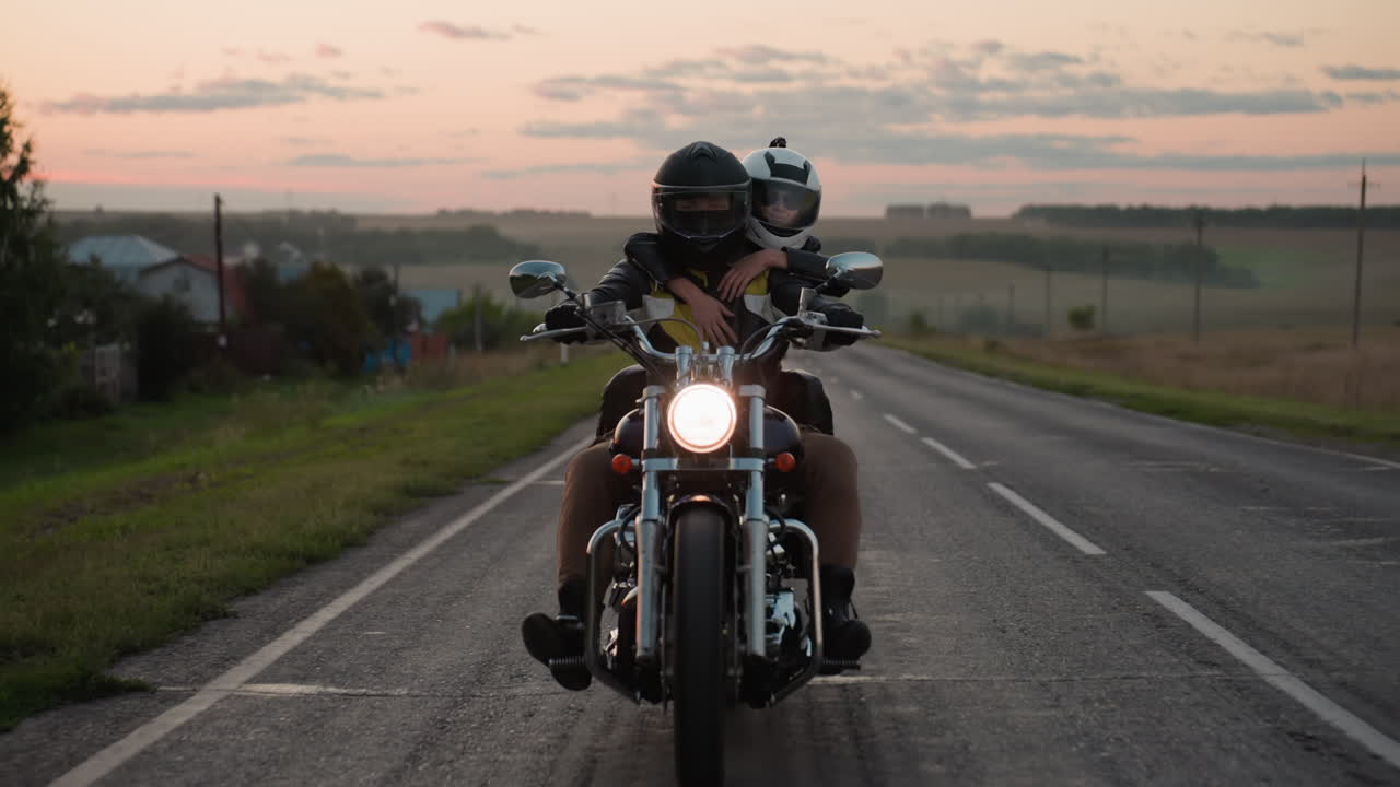 Two friends wearing helmets ride motorcycle on countryside road at sunset, passenger hugging rider tightly as glowing headlight lights up scenic path with rural fields and evening sky in background