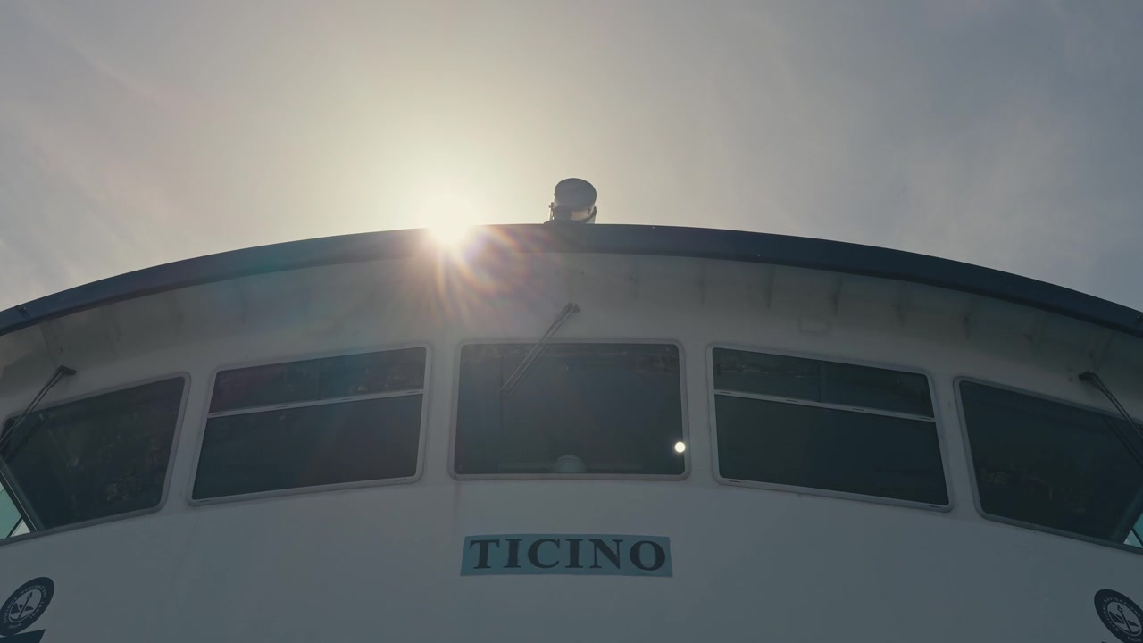 Primo piano medio che guarda in alto verso i finestrini del ponte di un traghetto passeggeri, con una luminosa stella solare che spunta dal tetto contro un cielo estivo limpido e blu.