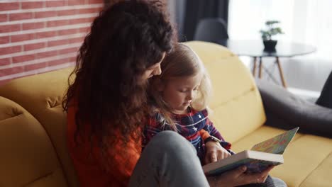 Kleines-Mädchen-Liest-Mit-Ihrer-Mutter-Ein-Buch,-Während-Sie-Zu-Hause-Auf-Dem-Sofa-Sitzt