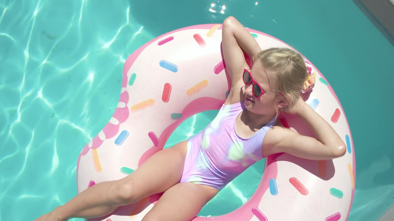 Girl child reclining on donut-float in pool, kicking, drifting to edge, fixing red shades, basking