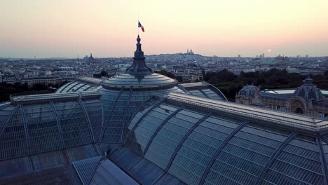 Aerial-panoramic-view-of-French-flag-waving-on-top-of-the-Grand-Palais-in-Paris-at-sunrise,-featuring-paris-city-and-Montmartre-at-distance