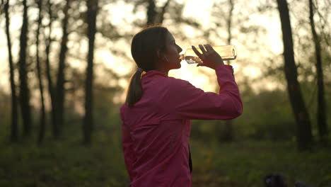 woman drinking water from a bottle in a forest at sunset