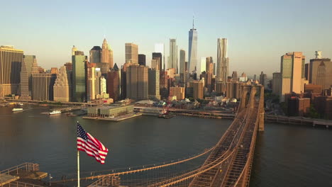 AERIAL:-Flight-backwards-over-Brooklyn-Bridge-with-Manhattan-view-close-up-of-American-Flag-at-Sunrise,-Sunset