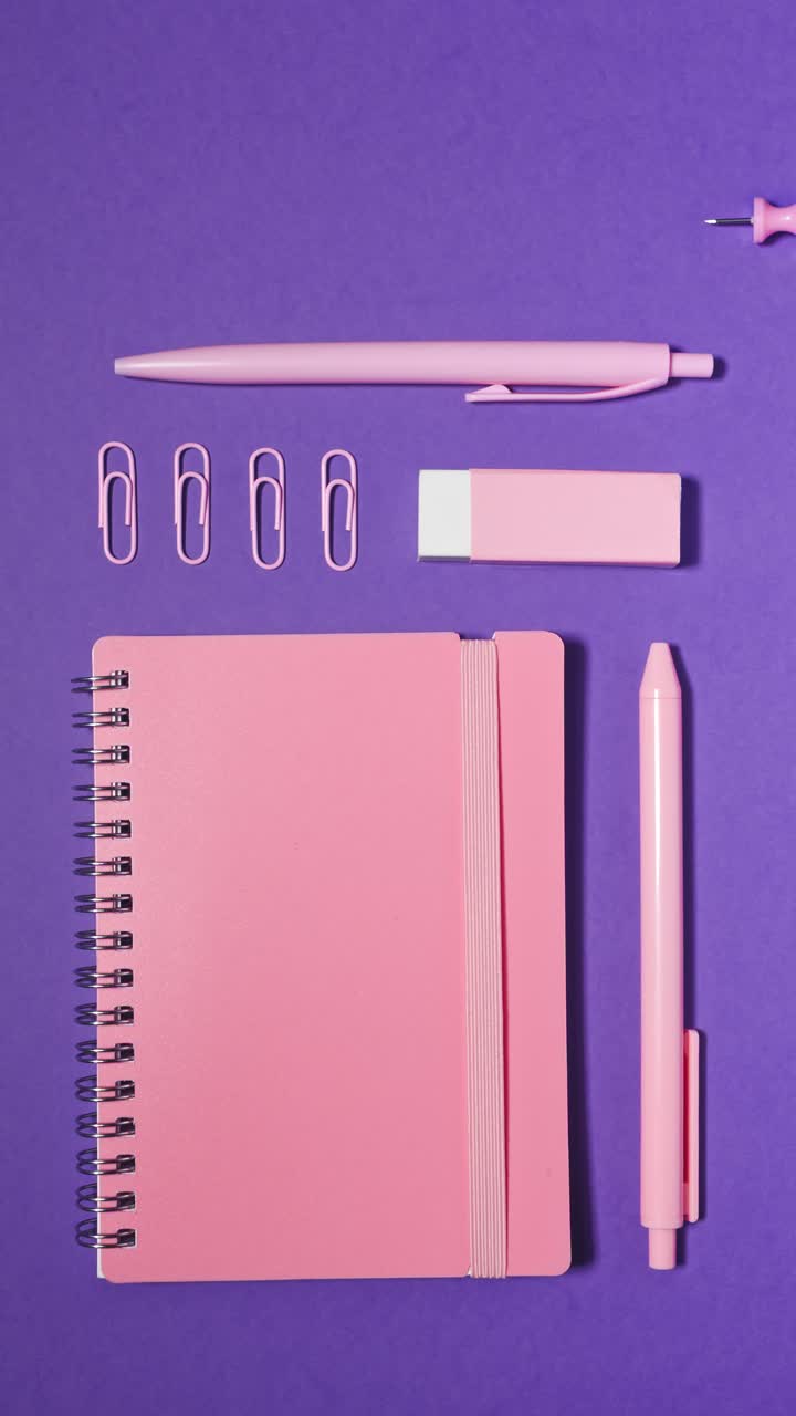 Vertical stop motion assembles pastel pink notebook, pens, clips, eraser and pushpins into tidy back-to-school layout on violet background