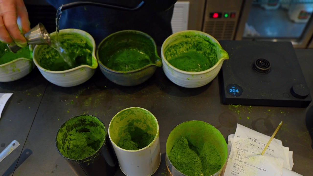 Chicago, USA, 27. maj 2026: Barista tilbereder matcha-drikke med skåle og kopper. Handskebeklædt hånd måler grønt matchapulver ved siden af keramiske skåle, papirkopper og en digitalvægt.