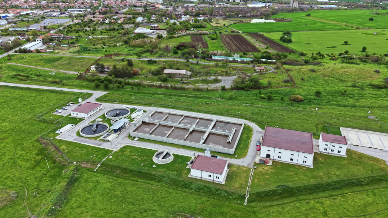 Large facility for water treatment located in a green area with fields and buildings visible in the background during a clear daytime
