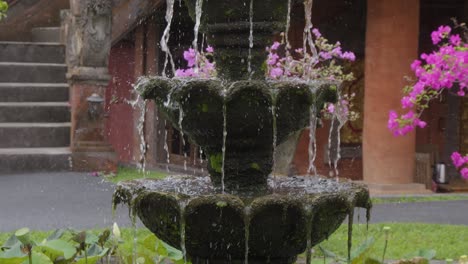 beautiful stone fountain with cascading water, surrounded by vibrant flowers, stands in front of traditional balinese architecture