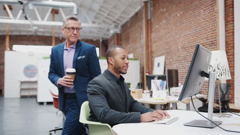 Mature-Businessman-Talking-To-Colleagues-As-They-Work-At-Desks-In-Modern-Open-Plan-Office