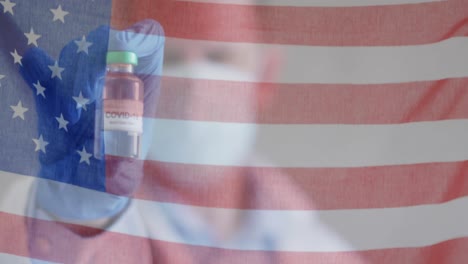 animation of american flag waving over male doctor in face mask holding covid 19 vaccine