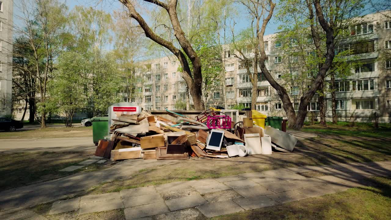 furniture, junk, electronics thrown on the ground near garbage bins in an Eastern European courtyard pov foward