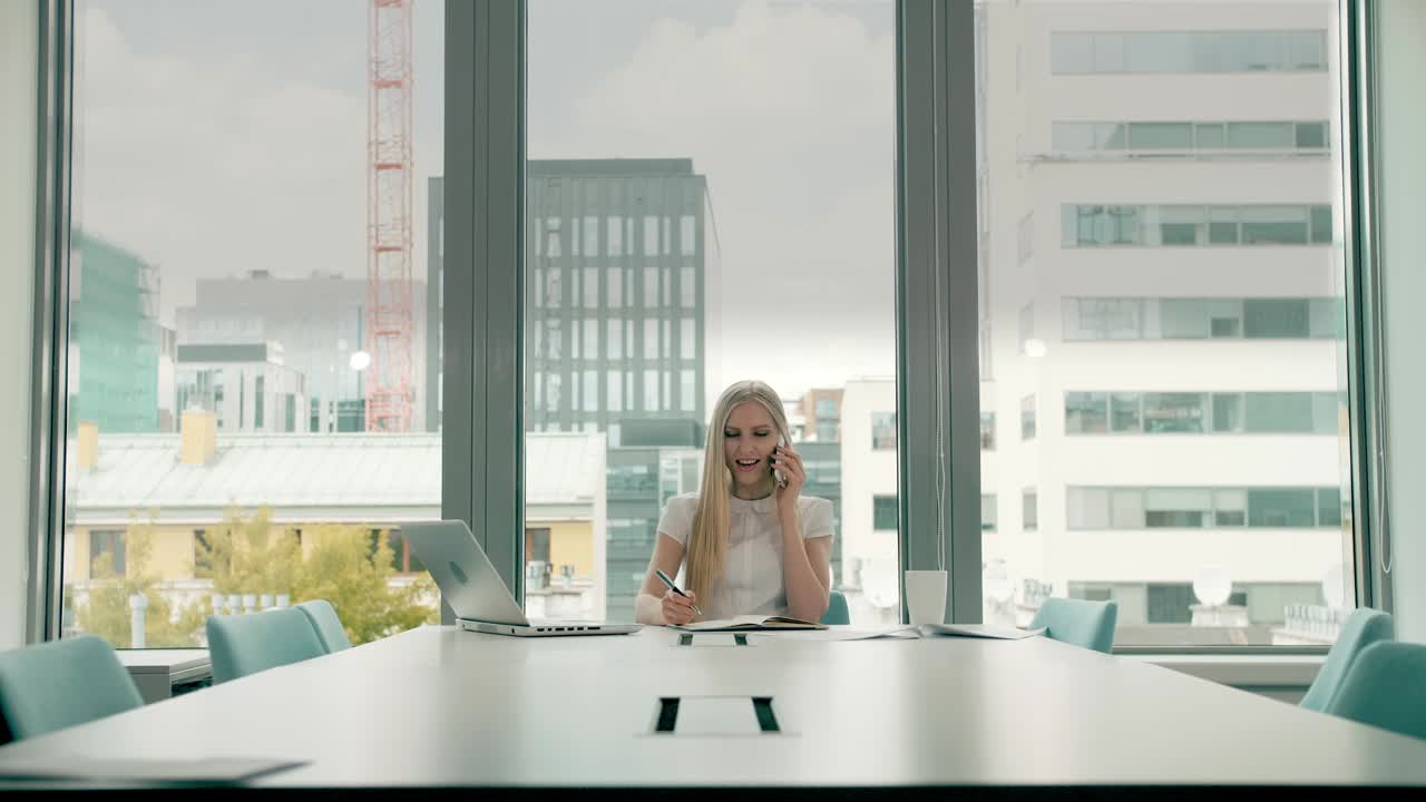 Businesswoman waving with hand while speaking on phone. Stylish businesswoman having phone call while sitting at table in conference hall and waving with hand greeting colleague