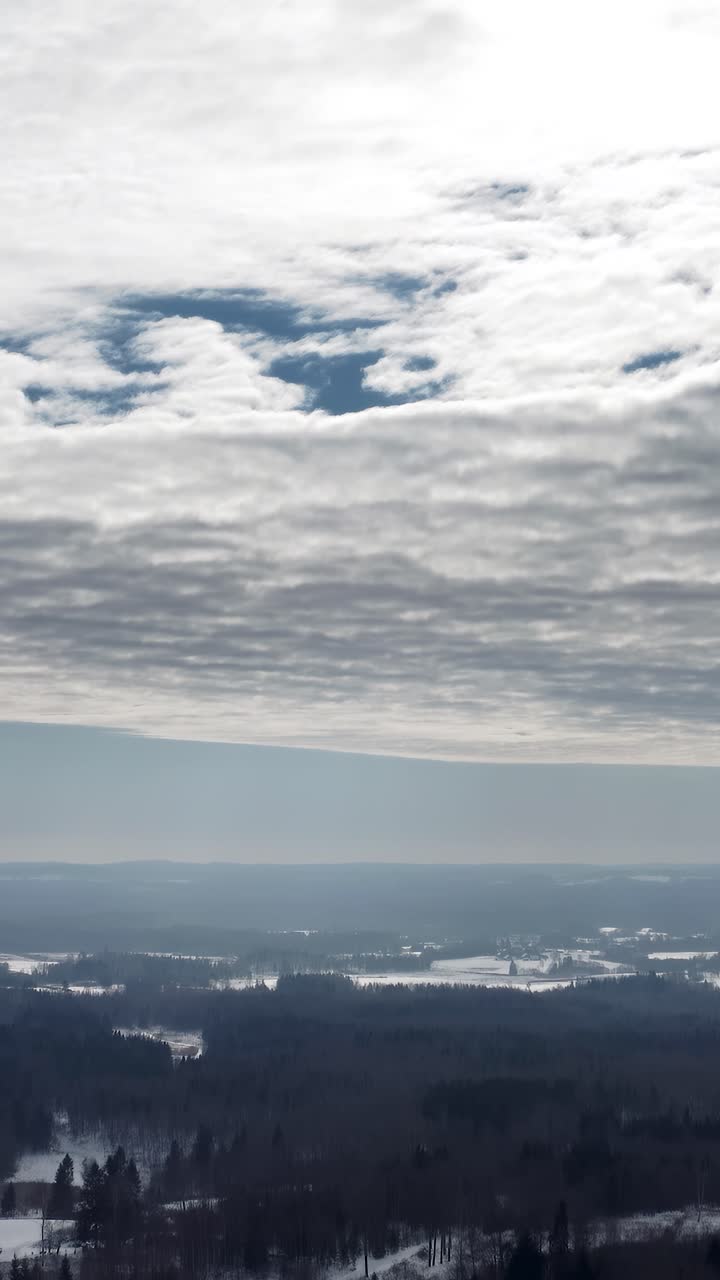 Vertical aerial drone view high up in the sky over a snow covered countryside landscape in winter. Sunrays through clouds over a dense forest.