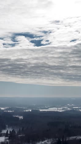 vertical aerial drone view high up in the sky over a snow covered countryside landscape in winter. sunrays through clouds over a dense forest.