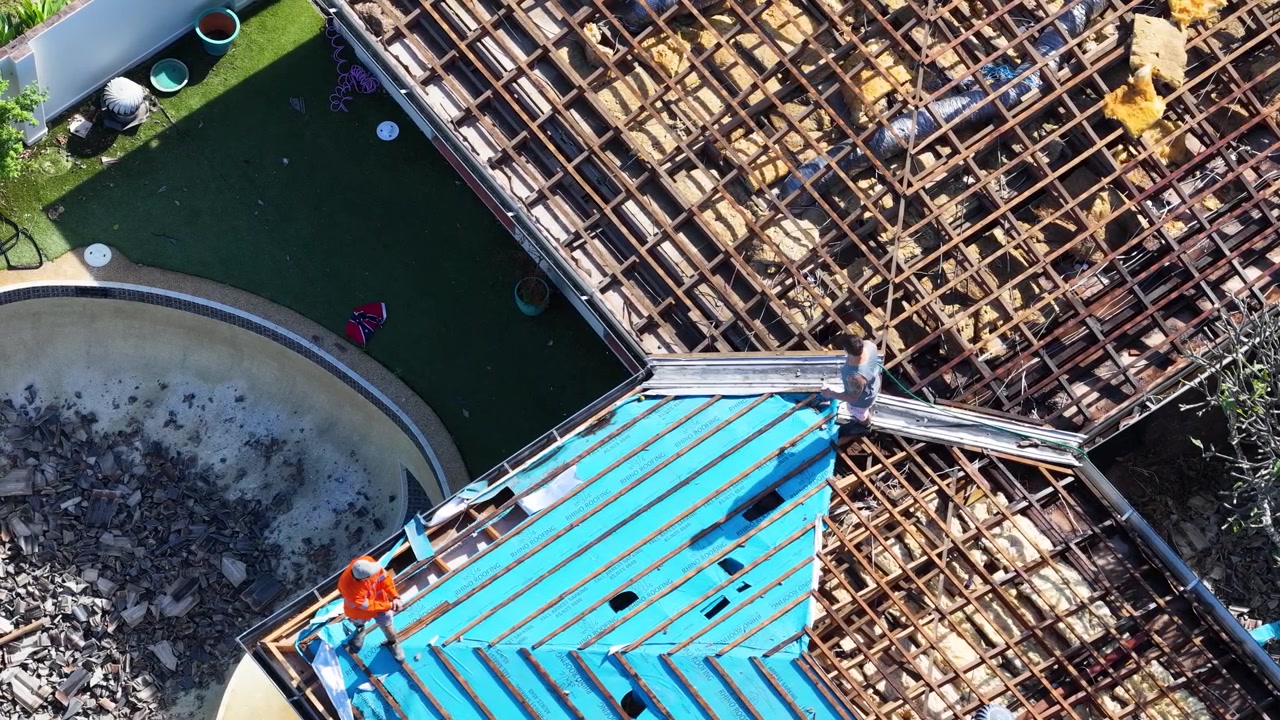 High angle drone shot shows laborers fixing house roof using blue insulation material under sunlight.