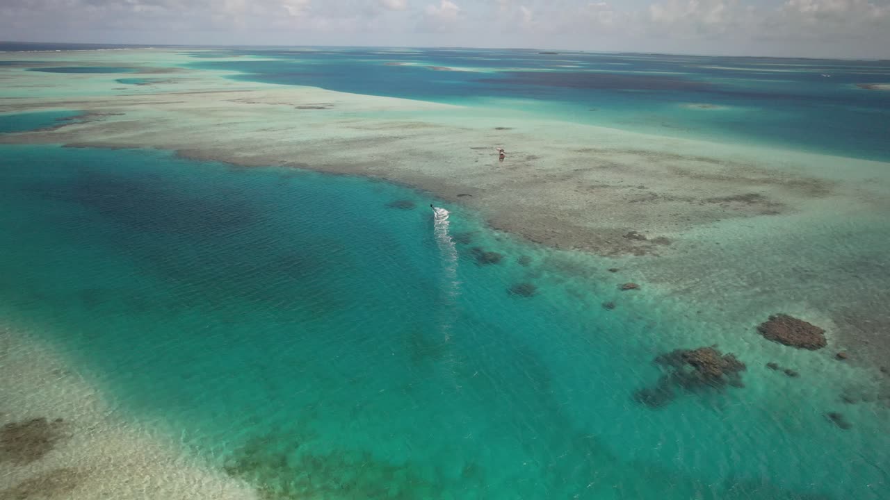 un kitesurfista solitario deslizándose sobre un mar azul de coral, alejándose de la cámara, vista aérea