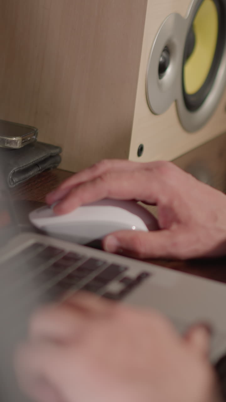 A close-up shot of hands working on a laptop, with one hand on the keyboard and the other on a mouse. The scene includes a blurred background and another person's hand resting on the table