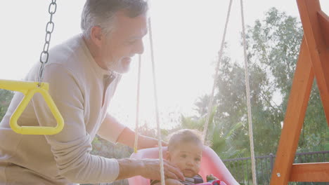 el abuelo hispano sonriente empujando a su nieto bebé en un columpio en un patio de recreo en el parque