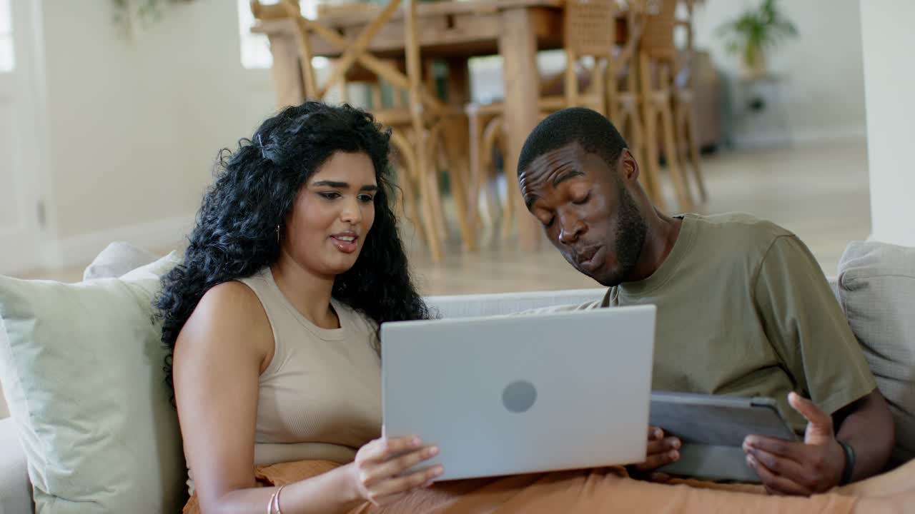 Couple with woman presenting laptop and man leaning from tablet viewing screen, smiling on sofa