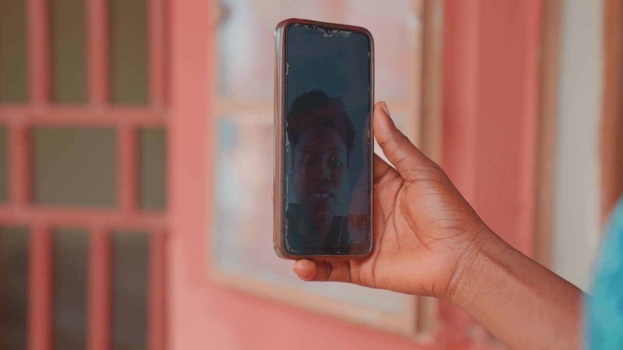 Videollamada de madre en smartphone, mano sostiene teléfono vertical con un rostro tenue en pantalla oscura, fondo cálido de hogar con ventana y pared pintada, momento íntimo de conexión entre madre e hijo/a, espontáneo.