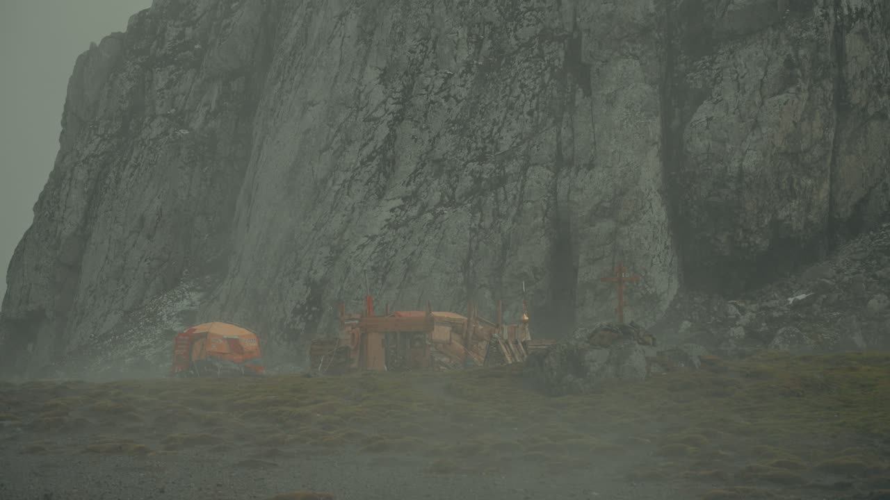 Base di ricerca russa improvvisata con rifugi arancioni raggruppati alla base di un'imponente scogliera a picco a Point Wild, Isola Elephant, a malapena visibile attraverso la nebbia geotermica vorticosa e il tempo rigido