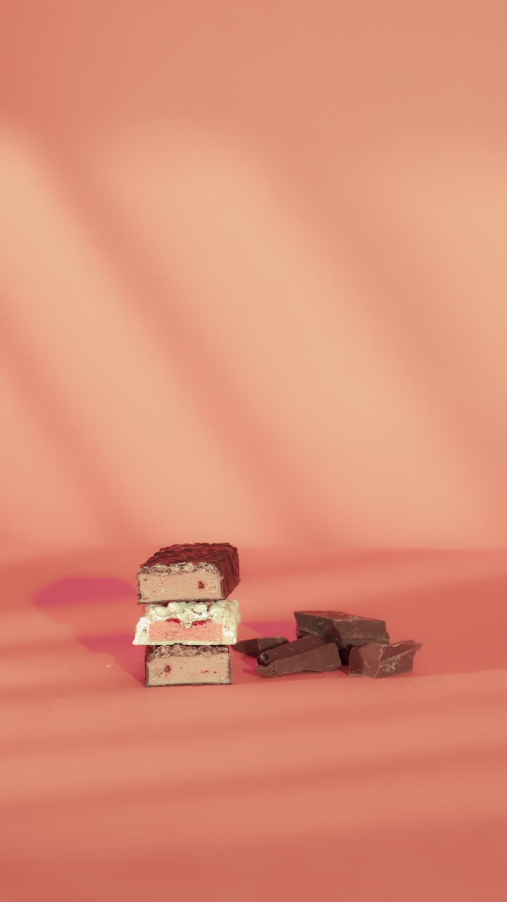 Close-up of a hand reaching to grab a stack of layered strawberry and chocolate bars on a pink surface beside broken chocolate pieces. Soft lighting highlights the textures and vibrant colors.