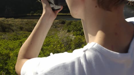 Young-boy-takes-camera-and-aims-to-majestic-coastline-to-take-photos,-close-up-motion-back-shot