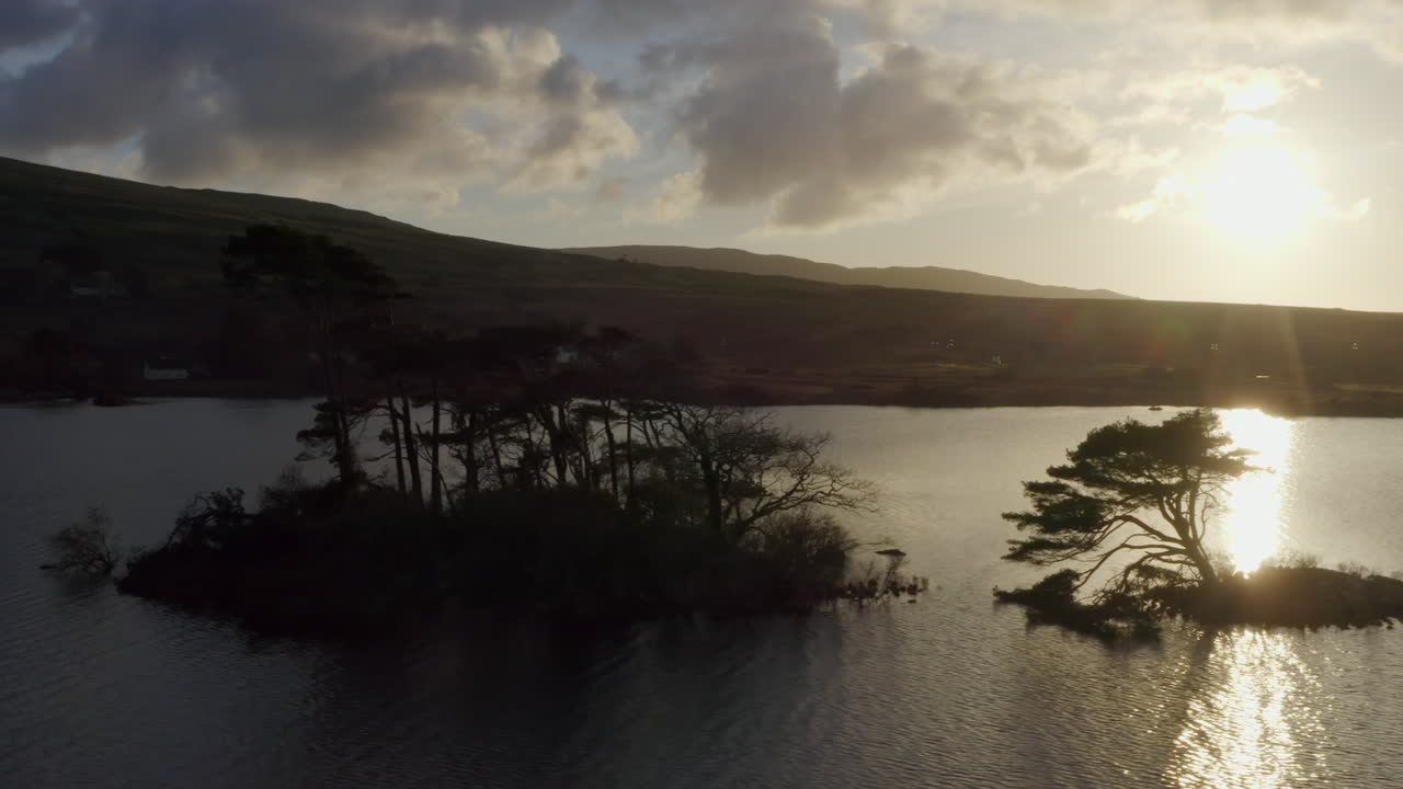 Aerial tracking left of islands backlit on Lough Bofin at golden hour sunset, the still lake waters defining the Irish landscape, Connemara, Galway, Ireland
