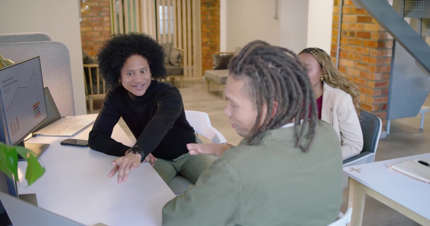 Team afroamericano che reagisce a un gesto con i dreadlock in riunione, mentre un uomo con dolcevita nero digita sul laptop.