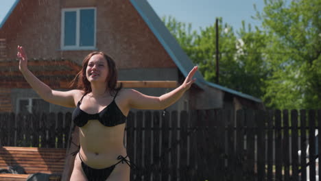 excited woman in black bikini jumps and laughs while playing under refreshing water spray in backyard, water droplets sparkle in sunlight as she embraces freedom and fun