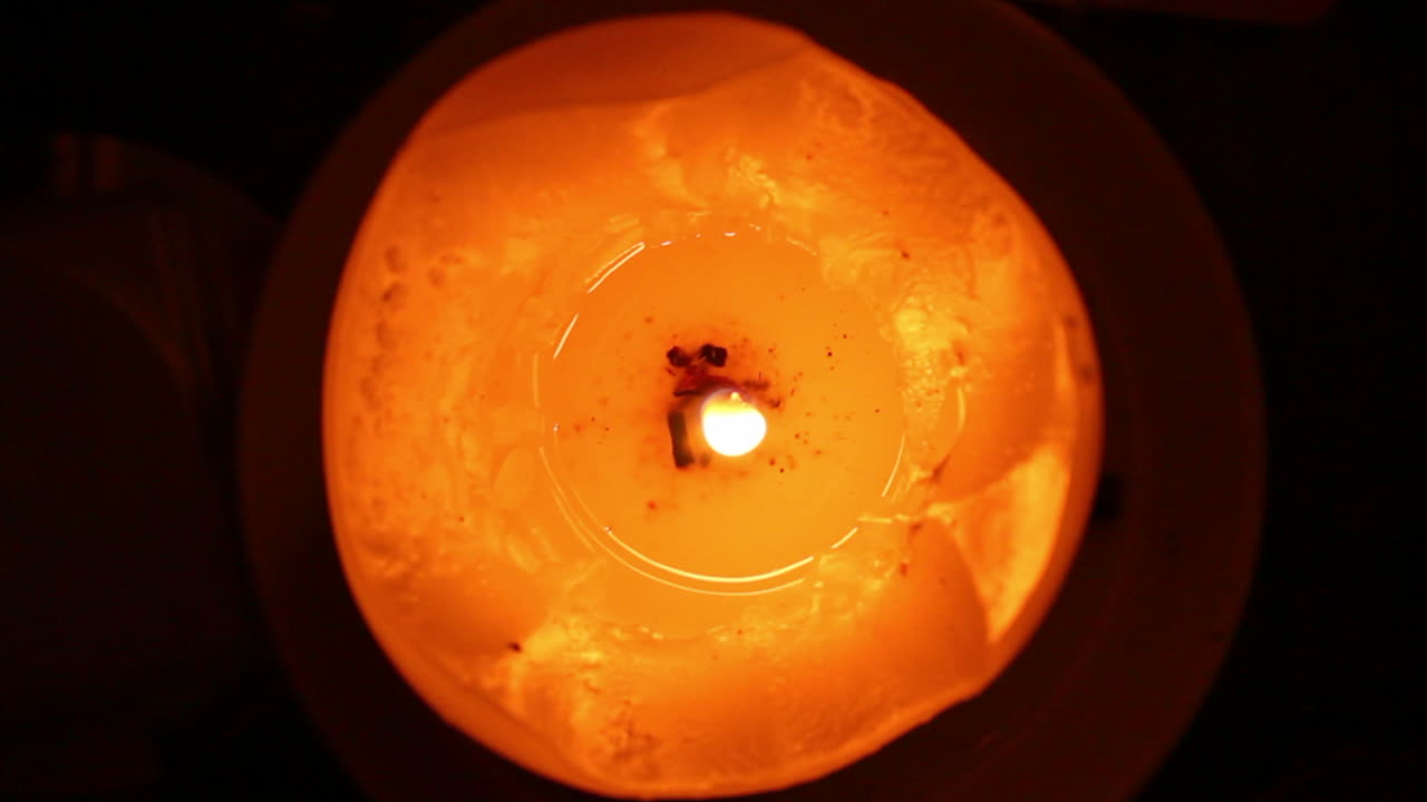 An orange and red candle with a litted shaky flame and melted wax on dark background viewed closely from above
