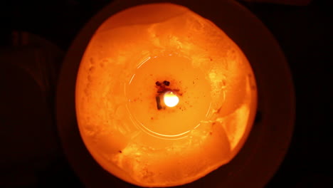 an orange and red candle with a litted shaky flame and melted wax on dark background viewed closely from above