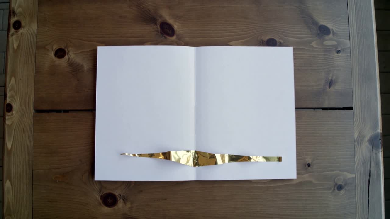 Il tempo che passa mostra una lamina dorata che si appiattisce, spargendosi lungo la rilegatura di un quaderno su un tavolo di legno.