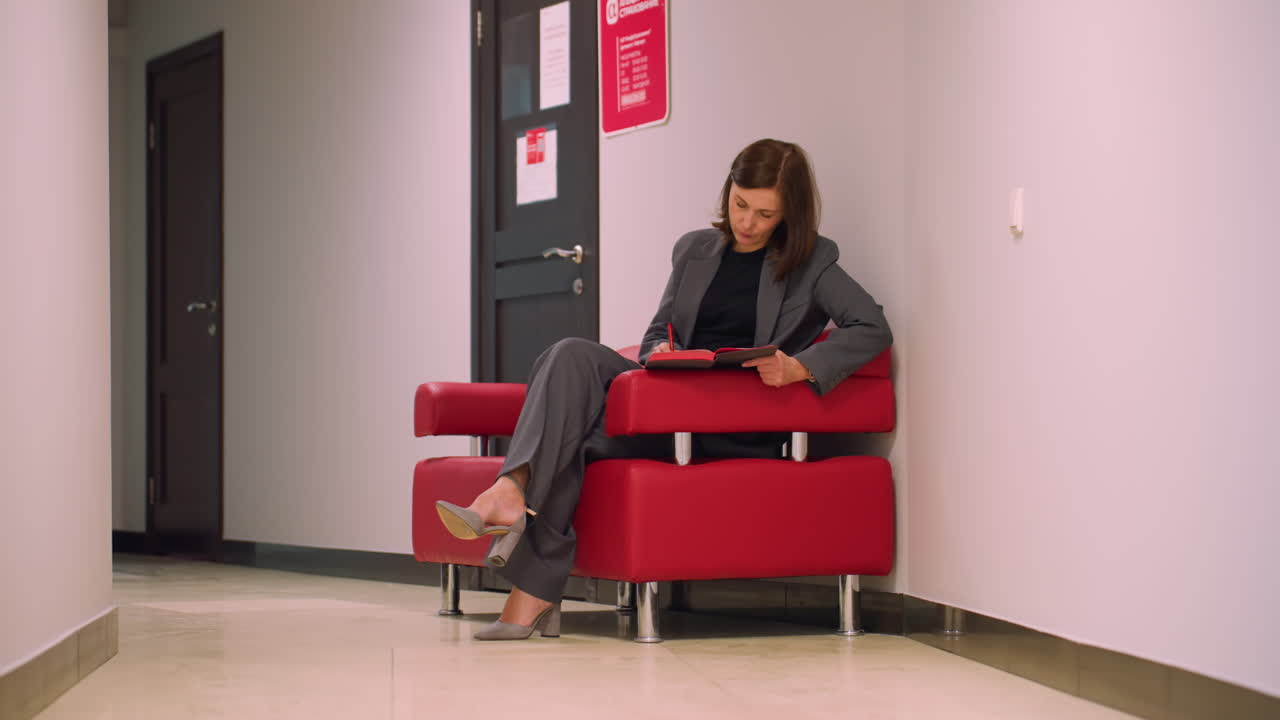 Businesswoman sitting on red chair in office hallway, writing in notebook with red pen, wearing gray suit and high heels, focused on work, calm professional setting, waiting for meeting