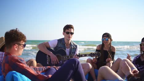 Young-man-playing-guitar-among-group-of-friends-sitting-on-easychairs-on-the-beach-and-singing-on-a-summer-evening.-Slowmotion-shot